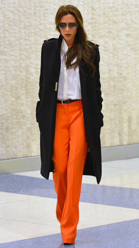 Victoria Beckham wearing orange pants, white top, and black trench coat