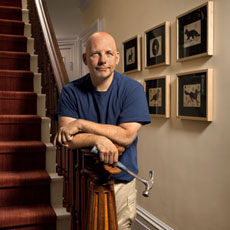 Scott Omelianuk leans on a baluster with hammer, framed posters in the background with inset of recent acquisition